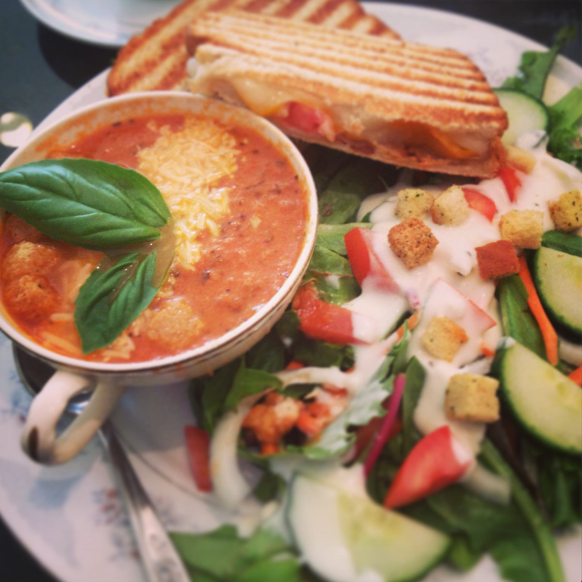 A close-up of a plated meal featuring a bowl of tomato soup with cheese and basil, a grilled sandwich, and a green salad with dressing, croutons, cucumber, and tomato on a plate.
