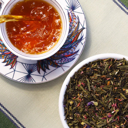 A top-down view of a white teacup and saucer filled with amber tea, alongside a white bowl of loose-leaf herbal tea blend on a patterned table mat.