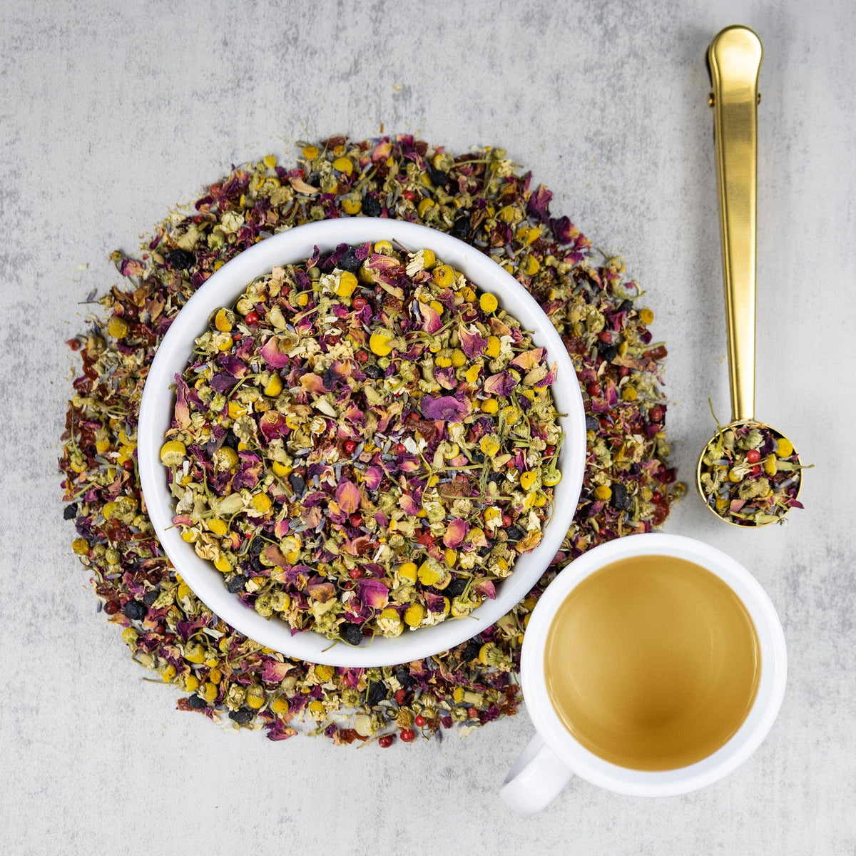 A top-down flat lay of loose-leaf herbal tea. A white bowl filled with colorful dried flower petals and herbs sits on top of a circular pile of the same tea blend, with a white cup of brewed tea and a gold measuring spoon on a light gray textured background.