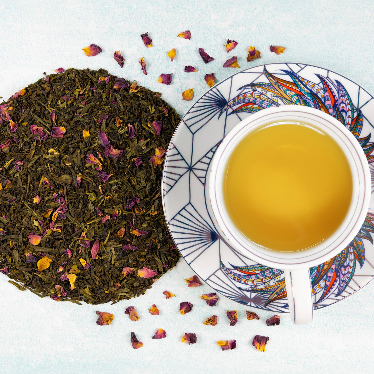 A top-down view of loose-leaf tea with dried rose petals, with one saucer holding a white cup filled with brewed golden tea, on a light blue textured background.
