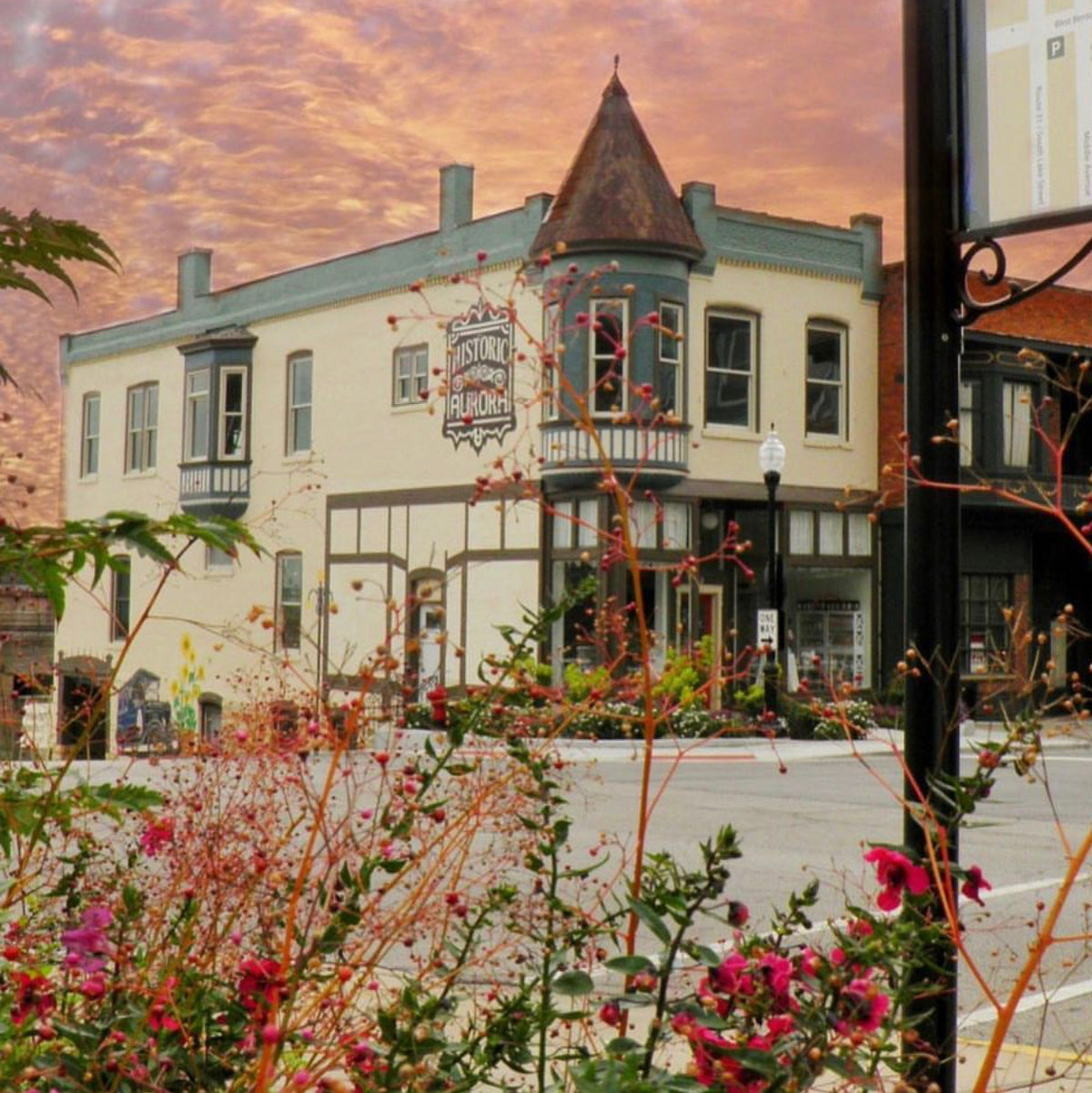 A scenic photograph of a historic cream-colored building with a conical tower, set against a dramatic orange and pink sunset sky. The foreground features blurred pink flowers and green foliage, framing the street scene.
