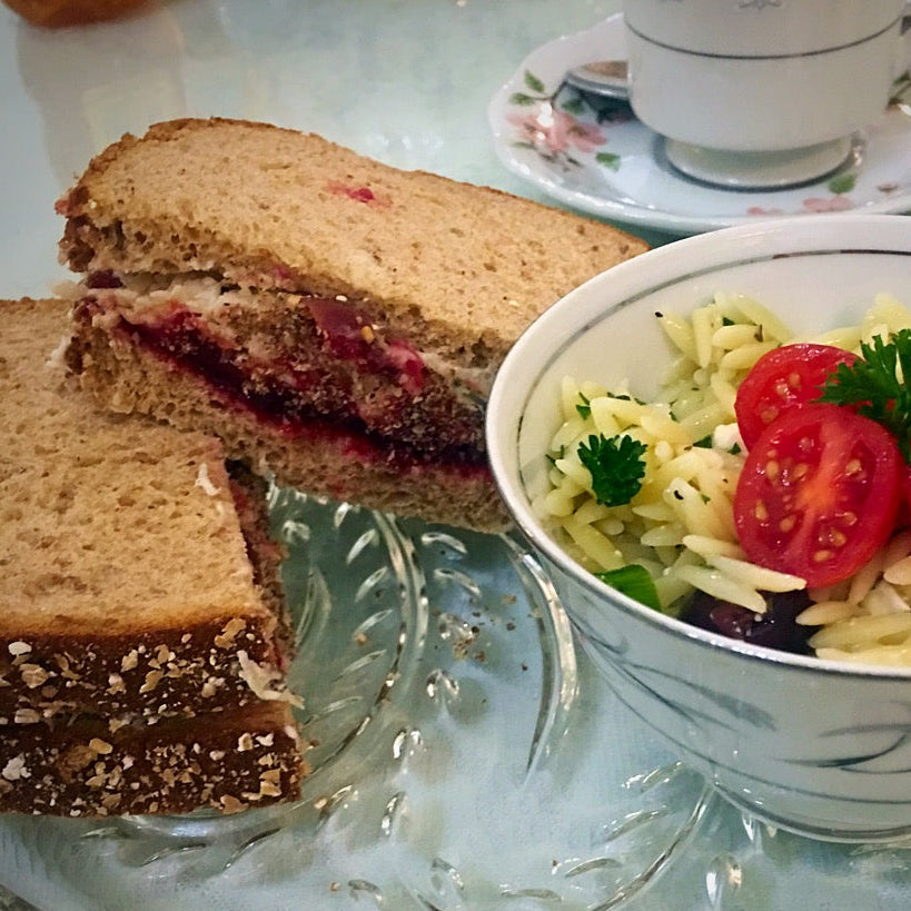 A close-up of a breakfast table setting featuring a sandwich with a dark berry jam filling on a clear glass plate, a bowl of orzo pasta salad with cherry tomatoes and parsley, and a floral teacup and saucer in the background.