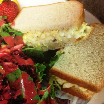 A close-up of a sandwich on white bread with a creamy yellow filling, served on a plate with a red salad and a strawberry.