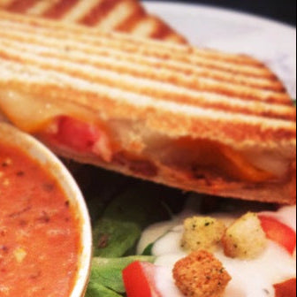 A close-up of a grilled sandwich on a white plate, served with a bowl of tomato soup and a side salad with croutons.