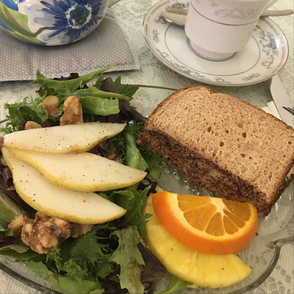 A close-up of a breakfast or tea-time spread on a table, featuring a glass plate with a mixed green salad, sliced pears, walnuts, an orange slice, and a slice of brown bread, alongside a floral teapot and a white teacup and saucer.