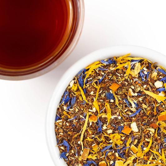 A close-up overhead shot of loose-leaf herbal tea in a white bowl, with a glass of dark amber tea at the top left. The tea blend contains brown tea leaves mixed with bright yellow, blue, and orange dried flower or citrus pieces on a white background.