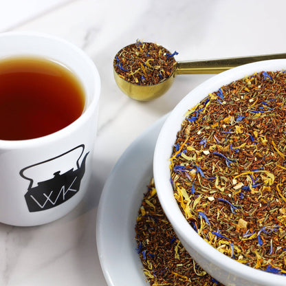 A close-up of a tea blend product shown in a white bowl and on a spoon, with brewed tea in a white mug beside it on a light marble surface. The tea contains visible dried flowers in yellow, blue, and brown tones.