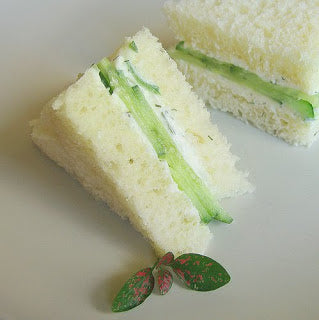 Two triangular sandwich slices filled with cucumber and white cream, arranged on a light beige surface with a small green leaf garnish.