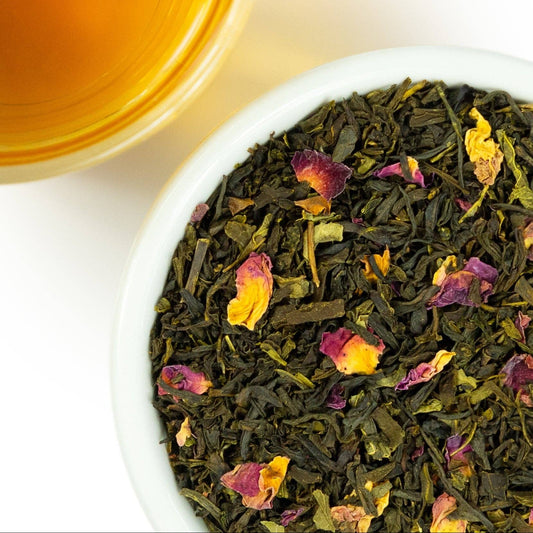 A close-up overhead shot of loose-leaf tea in a white bowl, with dried dark green leaves mixed with yellow and pink flower petals, next to a glass of amber tea on a white background.