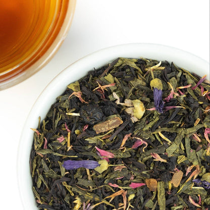 A close-up overhead shot of loose-leaf herbal tea in a white bowl, with a glass of amber tea in the upper left corner on a white background.